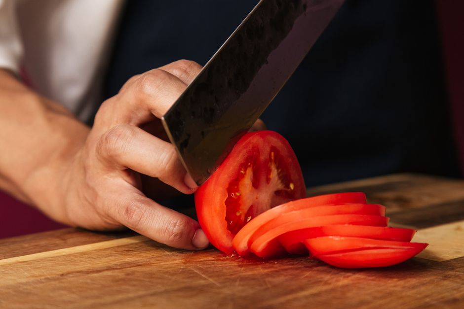 Chef's hand slicing ripe tomato on a wooden board with a knife. Perfect for food preparation visuals.